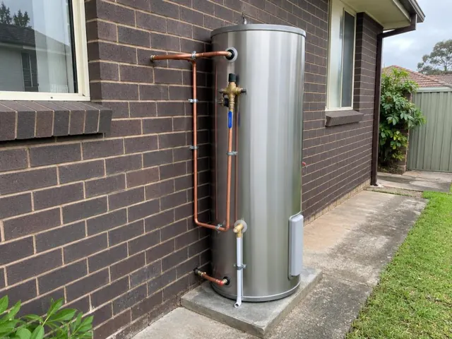 Outdoor stainless hot water tank with copper pipework beside a brick wall and concrete pad in Wentworthville
